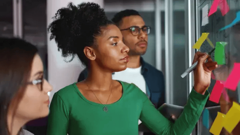 A black woman writing something on a white board with two people standing around her